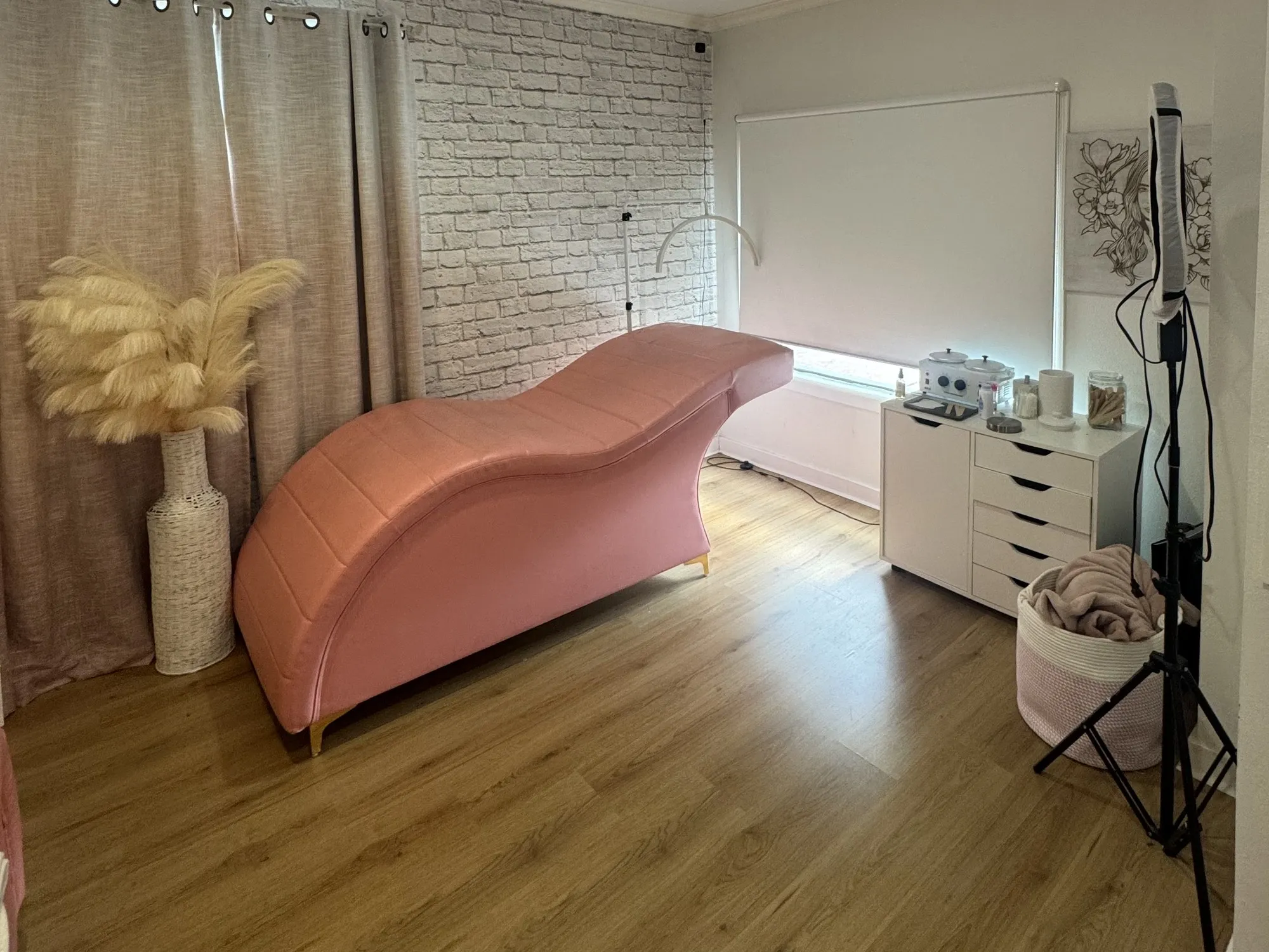 The room for rent at Eye Charm — a pink treatment bed, white brick wall, linen curtains and a window